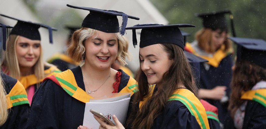 Two female students wearing cap and gowns look at a mobile phone screen.
